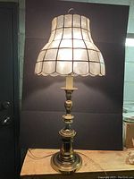 Full view of tall brass table lamp with shell shade on wooden table against black background.