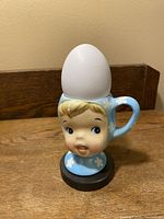 Front view of vintage Napco Miss Cutie Pie egg cup holding an egg, showing detailed facial features and blue hat with flower patterns.