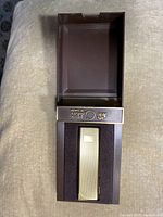 Top view of the gold-toned Colibri lighter inside its brown presentation box showing the lighter nestled within the molded interior.