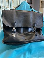 Front view of black leather messenger bag with flap closed showing two buckle straps and silver-tone hardware.