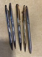 Five assorted pens arranged on a light surface showing two silver-tone pens with black tops and clips, one gold-tone pen, one gold-colored Cross pen, and one silver pen with texture.