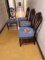 Four mahogany shield-back dining chairs in a row against a wall, showing embroidered blue seat cushions with floral designs.
