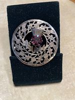 Front view of circular sterling silver brooch with detailed thistle pattern and central amethyst stone.