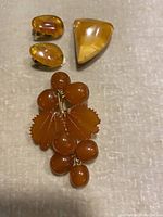 Top-down view of two brooches (amber grape cluster and triangular amber) and pair of amber clip-on earrings on a beige fabric surface.