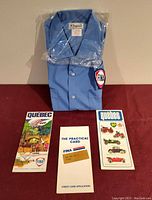 Folded blue FINA work shirt still sealed in plastic packaging with 2 Quebec road maps and a credit card application arranged in front