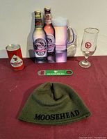 Photo of the 1970s Moosehead beer lot including cardboard beer bottle side, toque, bottle opener, beer glass, and empty beer can