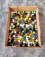 View of cardboard box filled with approximately 250 assorted vintage marbles in multiple colors and types with some visible play wear.