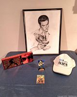 Overview of all items in the Elvis Presley lot, showing framed print, red chocolate tin, festival cap, figurine, and collectible card
