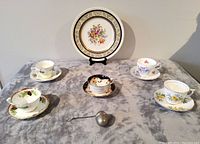 Full lot image showing five tea cup and saucer sets arranged around Johnson Bros plate and metal tea bag strainer in front