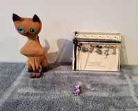 Wooden cat carving with blue eyes, pewter glass identifiers in original box, and pink jewelled cat pendant on grey surface.