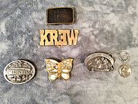 Six assorted brass and pewter belt buckles and keychains laid out on gray carpet, showing various designs including wildlife, letters, and commemorative firefighter motifs.