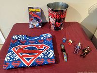 Full lot overview showing Superman sweatshirt, Spiderman book and tin bucket, small superhero figures, and Batman watch.