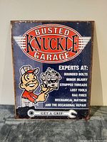 Front view of metal tin wall sign featuring Busted Knuckle Garage logo and humorous mechanic text on dark blue background.