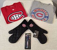 View of two Montreal Canadiens t-shirts (XL adult red and XL youth gray), two black quilted oven mitts with Canadiens logo, and NHL Canadiens pen in packaging on a flat surface.