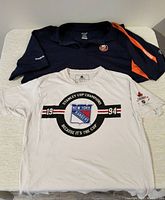 Two shirts displayed flat: New York Rangers white cotton t-shirt with front Stanley Cup Champion print and New York Islanders navy blue Reebok polo shirt with orange stripe on shoulder