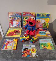 Lot of assorted Sesame Street Elmo books and toys displayed on a grey surface including plush doll, books, toy car and train