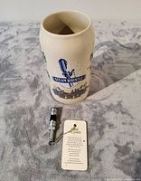 Ceramic beer stein with Steam Whistle logo and the 2018 pewter bottle opener with attached tag and chain on a gray fabric surface.