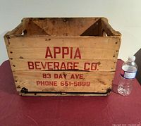 Front view of wooden crate with red stenciled text 'APPIA BEVERAGE CO., 83 DAY AVE., PHONE 651-5899' and plastic water bottle for scale.