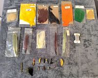 Top-down photo showing various packets of natural and dyed feathers and hackles, some still in original packaging, arranged in rows on a grey background.