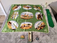 Green silk scarf with nine illustrated landmarks from Northern Ireland laid flat, displayed with various souvenir items including a tie, bell, pins, and spoon.