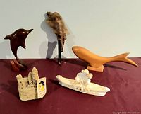 Five marine-themed vintage carvings arranged on a red cloth surface against a white wall. Items visible: wooden dolphin on base, driftwood flying fish, wooden fish, sandcastle sculpture with tag, salt sculpture of lovers on boat.