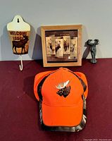 Photo showing the four items: orange cap with moose embroidery, wooden forest moose plaque, pewter moose figurine, and wall-mounted cup holder with 'Welcome to the Lodge' and moose image.