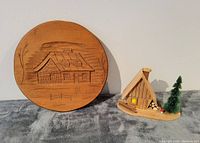 Photo showing the round carved wooden plaque with cabin scene and 3D wooden cabin model with tree, placed on gray fabric.