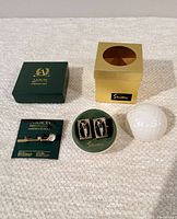 Overall view of two vintage golf-themed items: goldtone Stratton England cufflinks in original gold-tone box shaped like a golf ball container, abalone and Swarovski crystal goldtone lapel pin with display card, and a ceramic golf ball for scale.