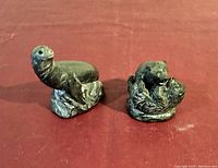 Front and side view of two dark soapstone animal carvings on red cloth: one seal, one pair of bear cubs playing, showing texture and shape details