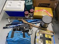 Photo showing assortment of camping and outdoor items including cooler, binoculars, umbrellas, thermoses, fishing rod, and trashcan with supplies.