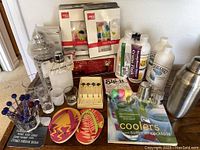 Photo shows various bar and drinking tools including plastic stirrers and drink picks, bottles for mixers, glassware, cocktail books, tropical coasters, and a metal cocktail shaker.