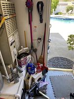 Vacuum cleaners, cleaning supplies, and household tools grouped near HVAC unit and doorway with pool visible outside