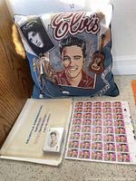 Elvis Presley pillow with colorful Elvis images beside a U.S. commemorative stamp album, a small box of Elvis-themed cards, and a sheet of 29-cent Elvis stamps.
