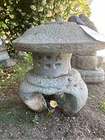 Close-up side view of the gray cement Asian pagoda garden statue with visible texture, windows, and curved legs, placed outdoors on soil with foliage background.
