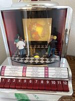 Front view of musical jukebox with 12 song selection buttons, dancing figurines on checkerboard floor, and projection on rear screen.