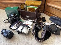 Wide shot showing Olympus OM-10 camera with lens and original box, Fujica and Minolta cameras, Vivitar flash, leather camera case, straps, and additional lenses scattered on carpet