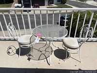 Full view of the patio set showing round glass-top table and two matching chairs with white painted metal frames and vinyl cushions.