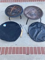 Two outdoor portable metal fire pits side by side with mesh covers, foldable legs, fire pokers, and fabric carrying covers on a concrete ground with brick edging.