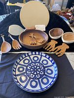 Overall view of all items in the lot: Pampered Chef baking stone, ceramic serving bowl, wooden bowls and utensils displayed on black cloth.