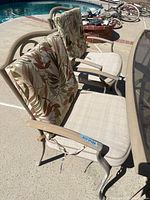 Side view of metal patio chairs featuring armrests and leaf-patterned cushions on backrest and beige fabric seat cushions.