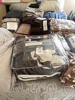 View of multiple soft throws and blankets in neutral tones stacked on bed alongside packaged bedding set.
