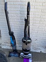 Two upright vacuums side by side, Shark Navigator (black/silver) and Bissell blue cordless multi-surface vacuum