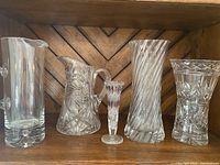 Full group shot showing all five crystal and glass items on a wooden shelf: two pitchers, three vases with various cut patterns and designs.
