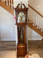 Full view of the oak-finish wooden clock standing 74 inches tall against a wall and staircase.