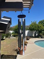 Metal and wood garden wind chimes hanging outdoors near a pool and plants, shown in bright daylight from a frontal perspective.