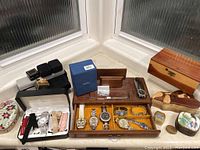 Overhead view of multiple women's watches arranged in wooden and fabric watch boxes with additional jewelry boxes and small trinkets around.