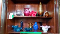 Full view of wooden cabinet shelf holding various art glass items featuring vases, bowls, and pitchers.