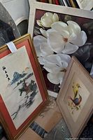 Photo showing three framed artworks leaning against a wall: one floral print, one bird painting, and one Asian embroidery landscape.