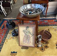 Photo shows a framed vintage Asian puppet in a shadow box resting against a wooden stool. To the left is a black metal vintage lamp and to the right a woven basket and a silver metal lamp with cord on the floor around the stool, which holds a large metal bowl filled with assorted kitchen cutlery.