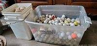 Wide view of two plastic totes; one white tote containing used baseballs and a larger clear tote filled with used golf balls.